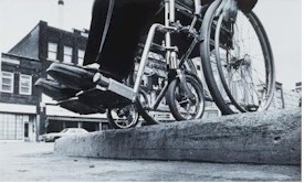 Wheelchair at the edge of an 
      un-cut curb. Select for more details of the picture.