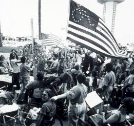 Picture of demonstrators with ADAPT flag and chair hanging from cross. Select for more details.